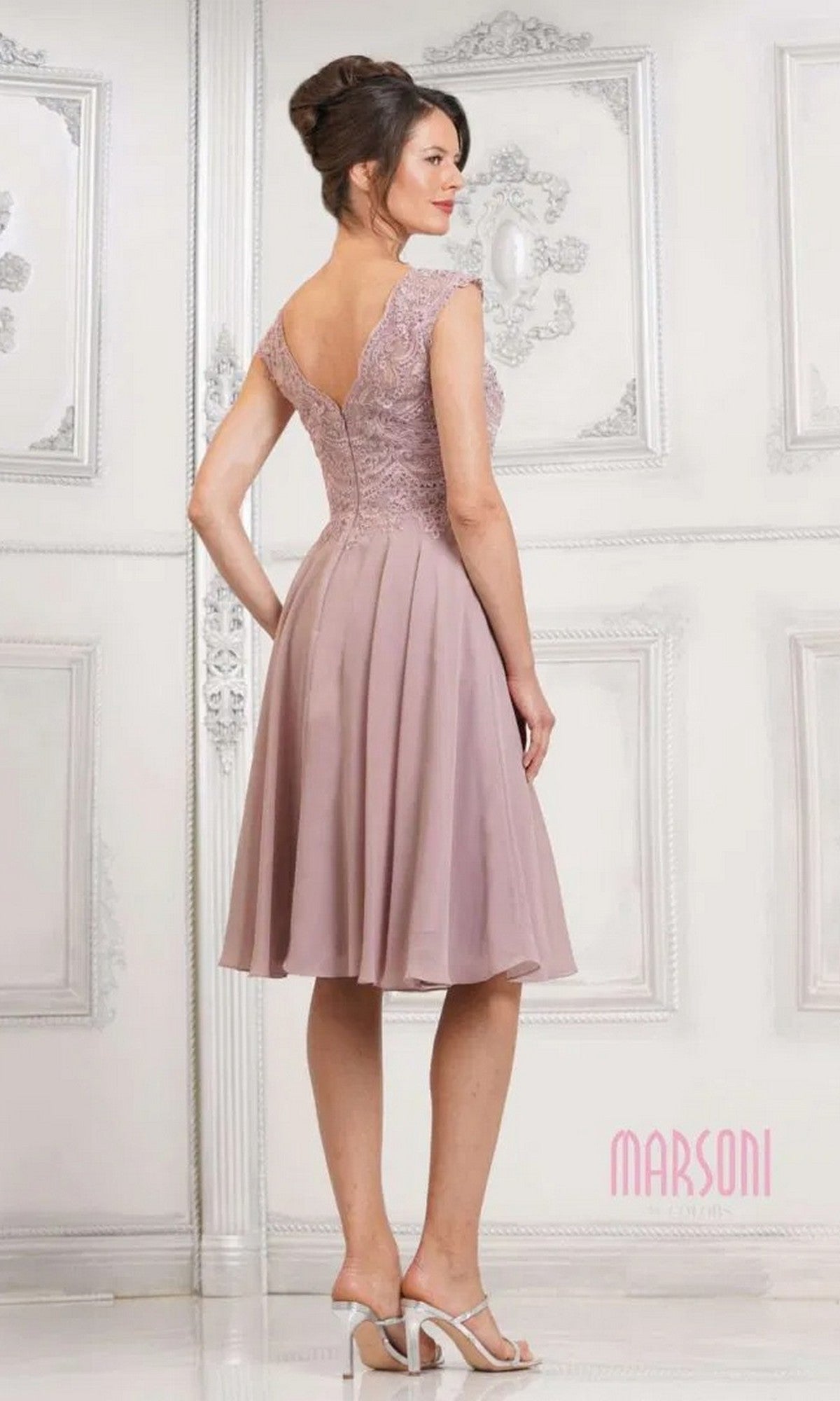 A woman faces away, wearing the Marsoni M238S knee-length semi-formal dress in mauve, with a lace-embroidered bodice, chiffon skirt, V-back, cap sleeves, and paired with white heeled sandals.
